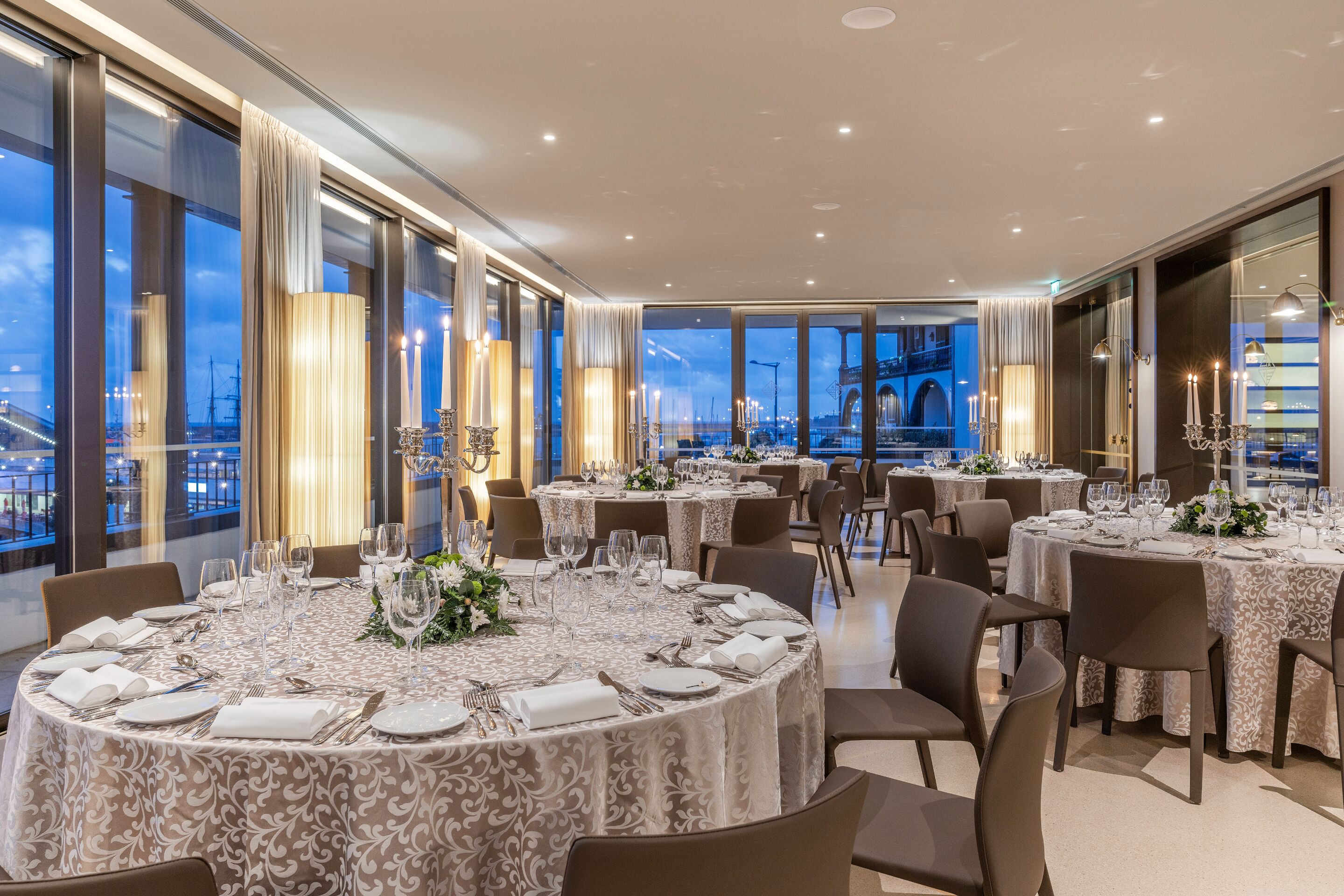 Un restaurante de lujo con mesas redondas dispuestas para comer, amplias ventanas que ofrecen vistas a la ciudad y una elegante vajilla.