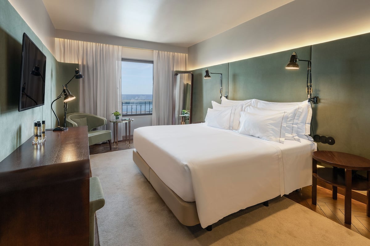 Een hotelkamer met een groot bed, een bureau met lamp, een stoel, een klein tafeltje en uitzicht op zee door het raam.