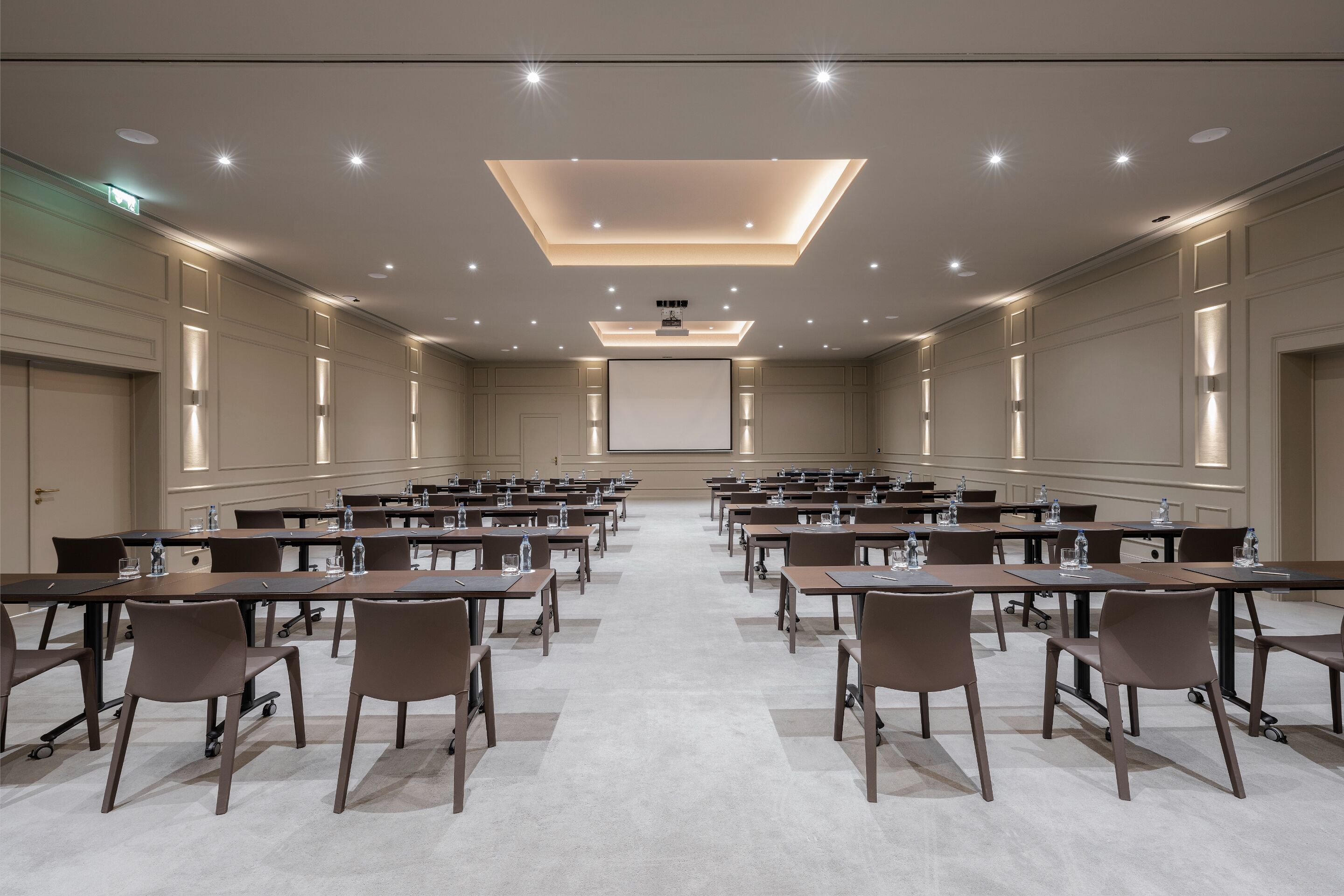 A conference room with rows of tables and chairs, a large screen at the front, and recessed ceiling lighting.