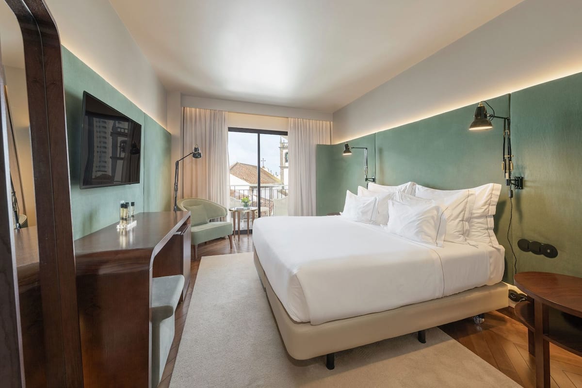 Een hotelkamer met een bed, bureau, stoel en uitzicht op een gebouw met een toren.