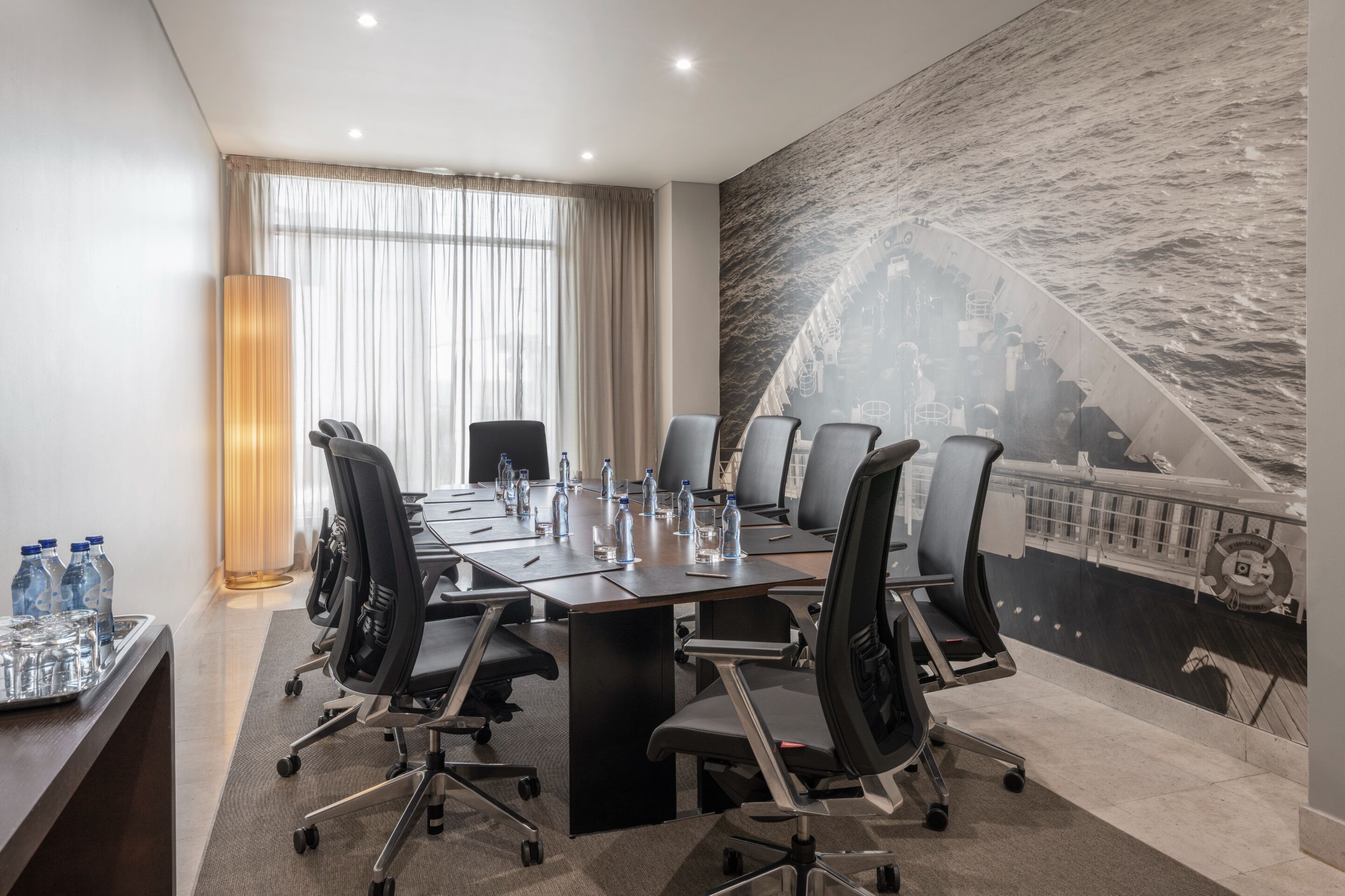 A conference room with a long table, multiple chairs, water bottles, and glasses. There is a large window with curtains and a mural of a ship on the wall.