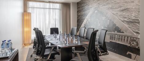 A conference room with a long table, multiple chairs, water bottles, and glasses. There is a large window with curtains and a mural of a ship on the wall.