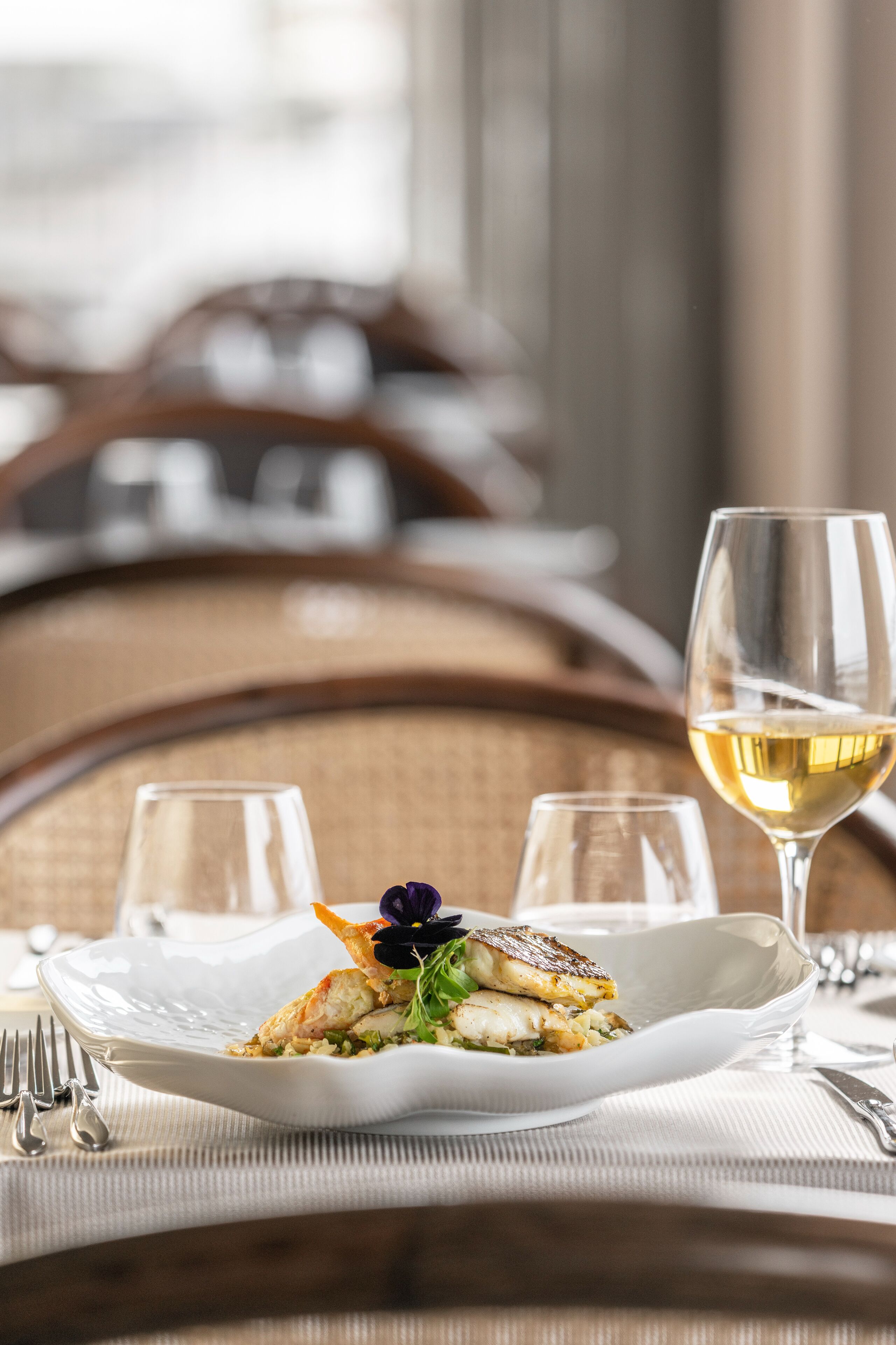 A dining table with a white plate of food, a glass of white wine, and a fork.