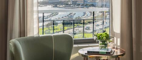 A room with a view of a cityscape through a window, a green upholstered chair, and a small table with a vase of flowers.