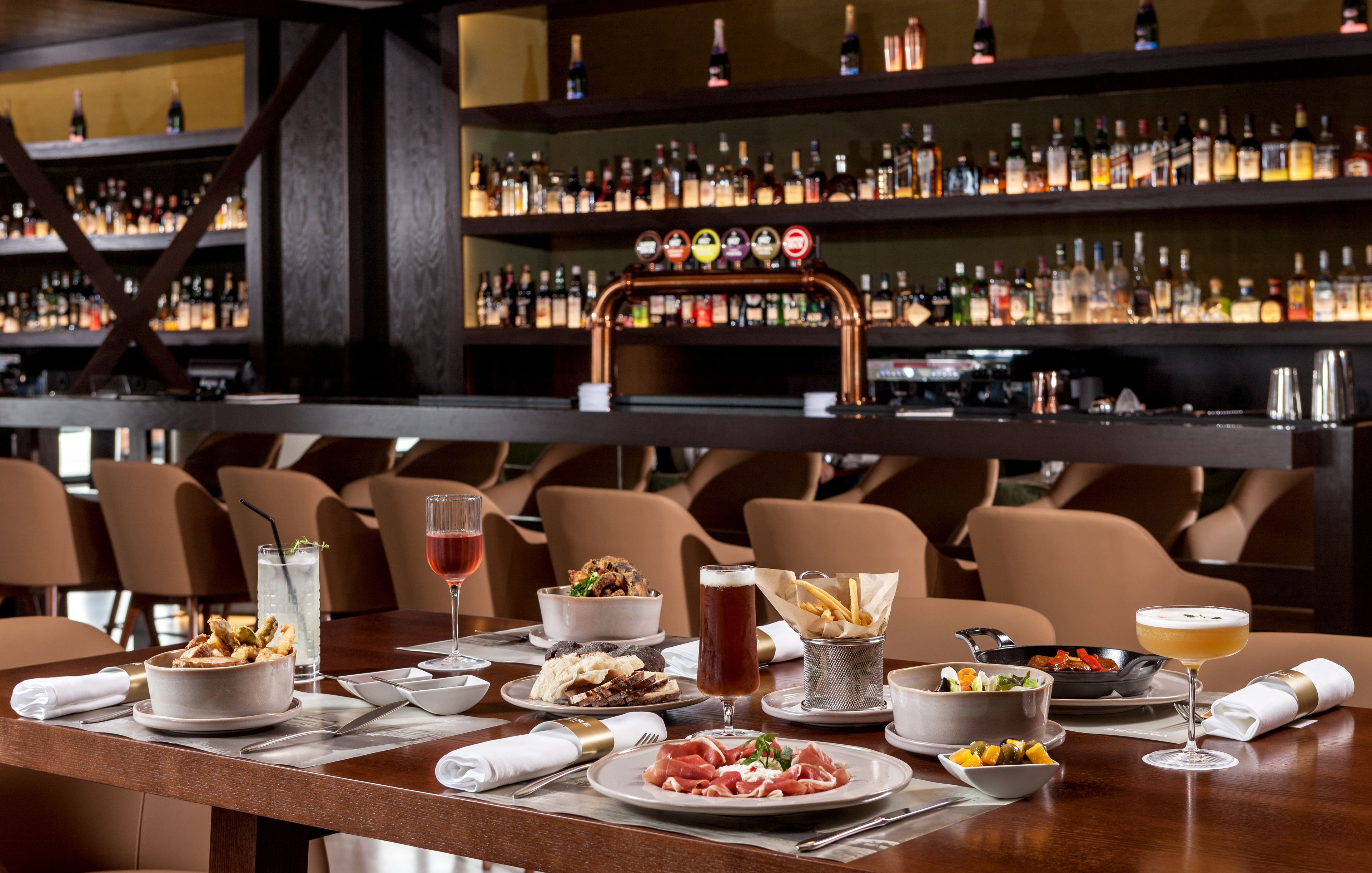 Uma área de bar com várias prateleiras de garrafas, uma torneira de cobre e uma variedade de pratos e bebidas sobre a mesa.