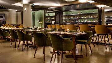 Un restaurante con sillas de terciopelo verde, mesas de madera y una zona de bar con estantes llenos de botellas.
