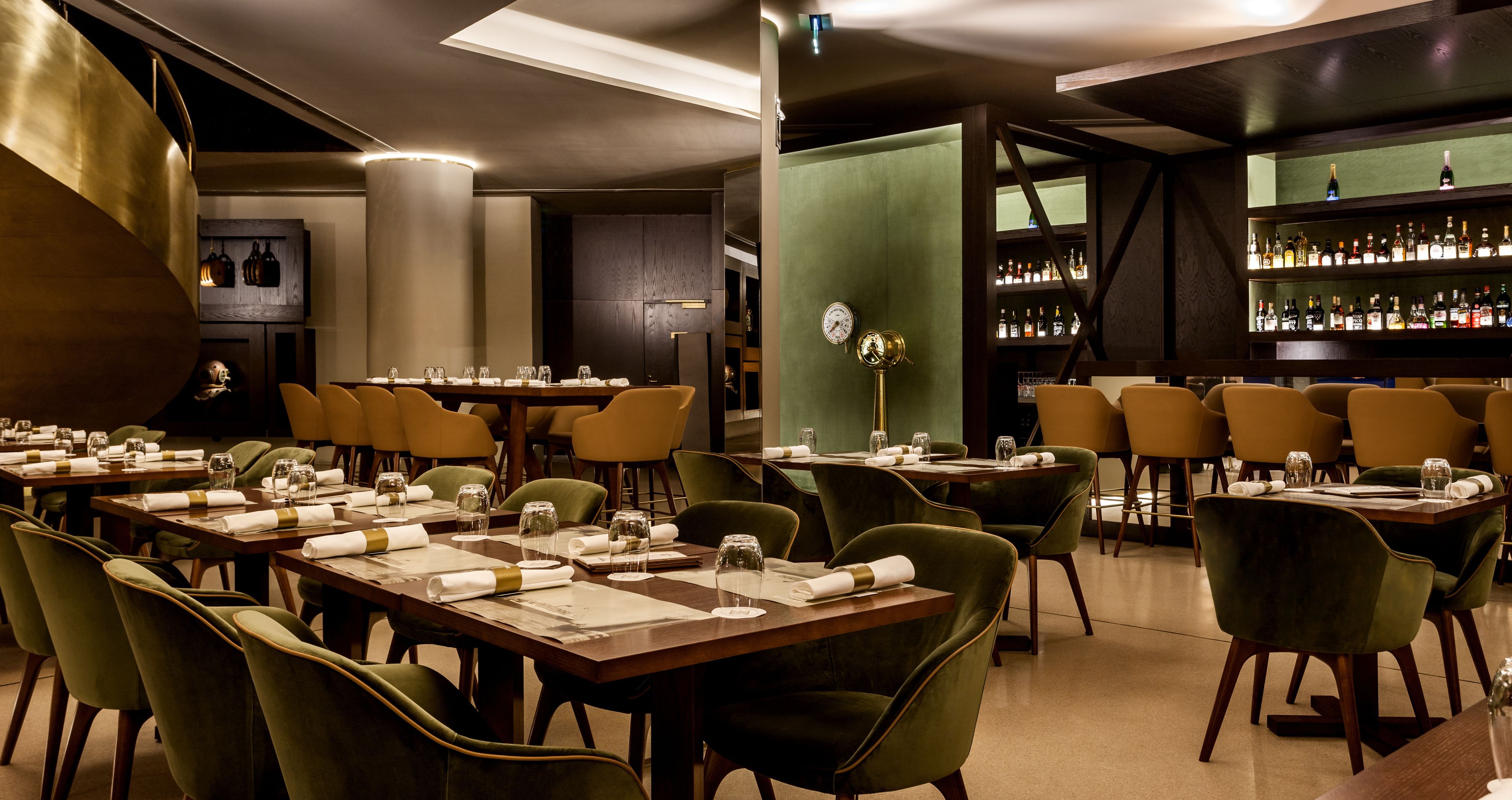 En restaurant med flere borde dækket til spisning, et barområde med hylder fyldt med flasker, og en stor dekorativ metalstruktur.