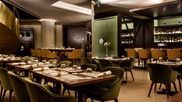 En restaurant med flere borde dækket til spisning, et barområde med hylder fyldt med flasker, og en stor dekorativ metalstruktur.