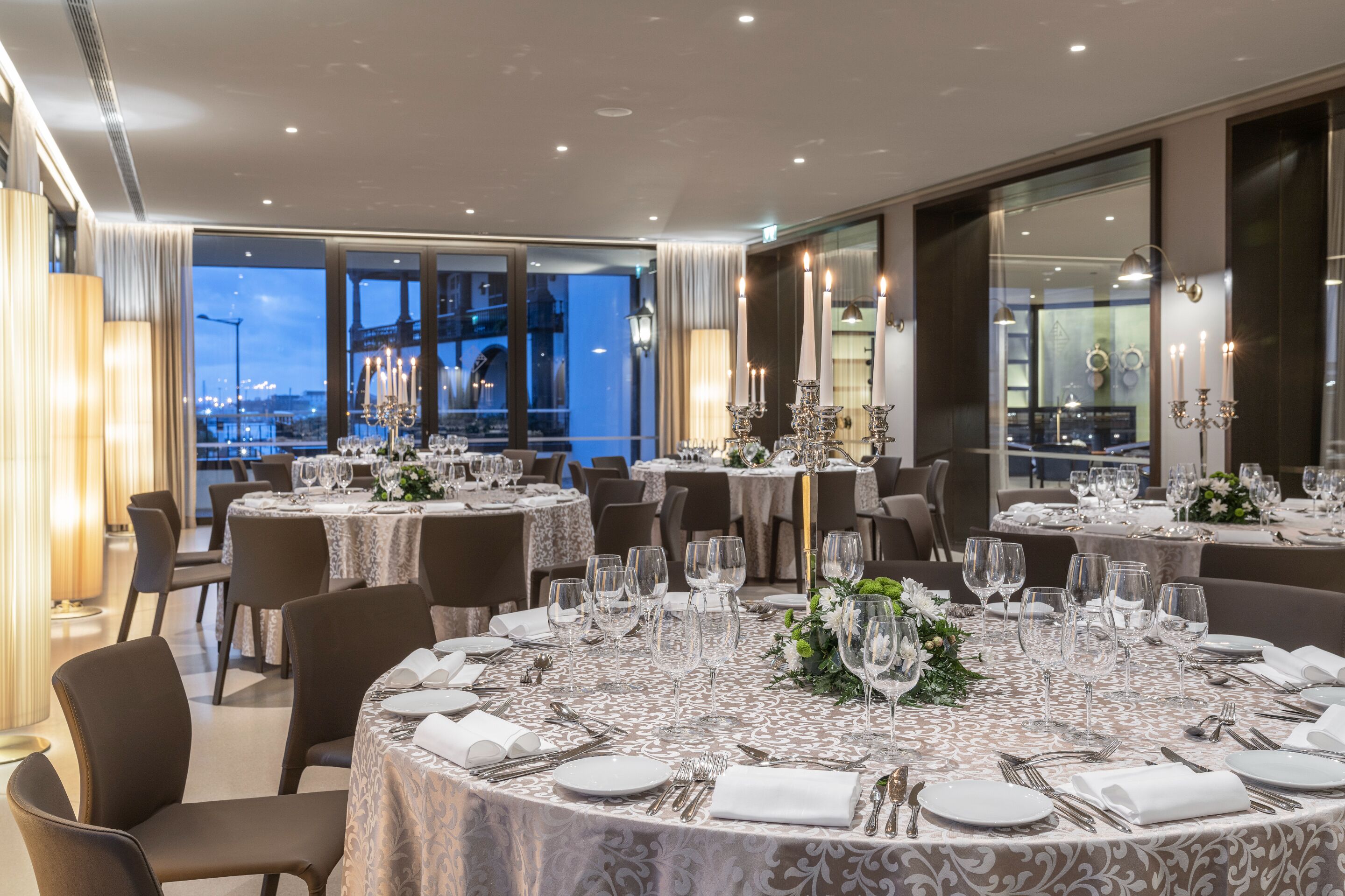 Comedor con mesas redondas dispuestas para una comida formal, con manteles blancos y cubertería.