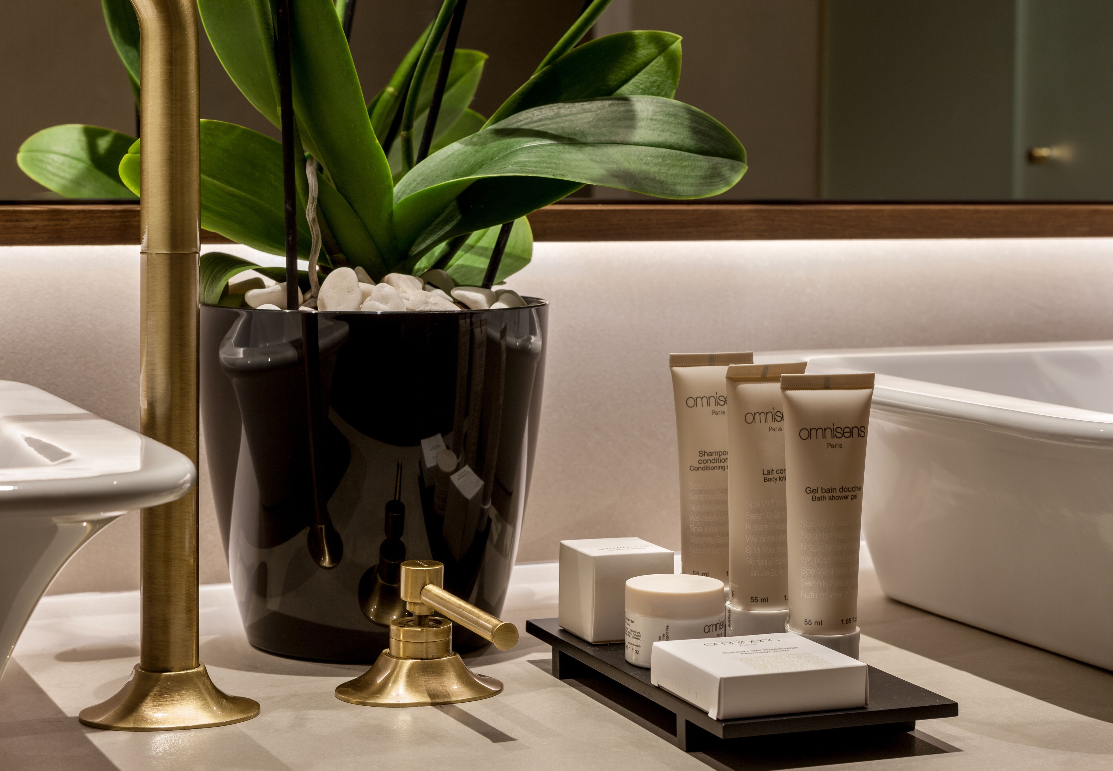 Un baño con un florero negro que contiene guijarros blancos y hojas verdes, un grifo dorado y varios artículos de tocador, incluyendo una botella de champú y un tubo de crema corporal.