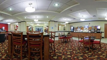 A hotel dining area with multiple tables and chairs, a central counter with a coffee machine, and a patterned carpet.
