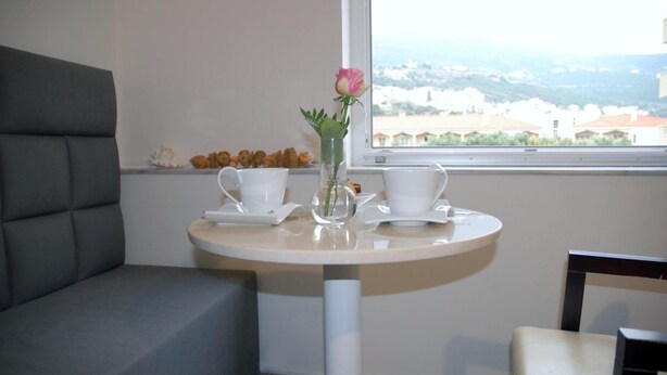 A small round table with two white cups and saucers, a vase with a pink flower, and a window with a view of buildings. - Hotel Aria (Samos)