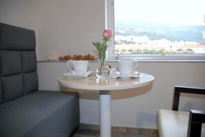 Una pequeña mesa redonda con dos tazas y platillos blancos, un florero con una flor rosa, y una ventana que ofrece vistas a edificios.