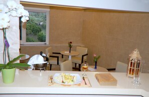 A dining area with a table set for two, a vase with a flower, and a view of greenery outside.