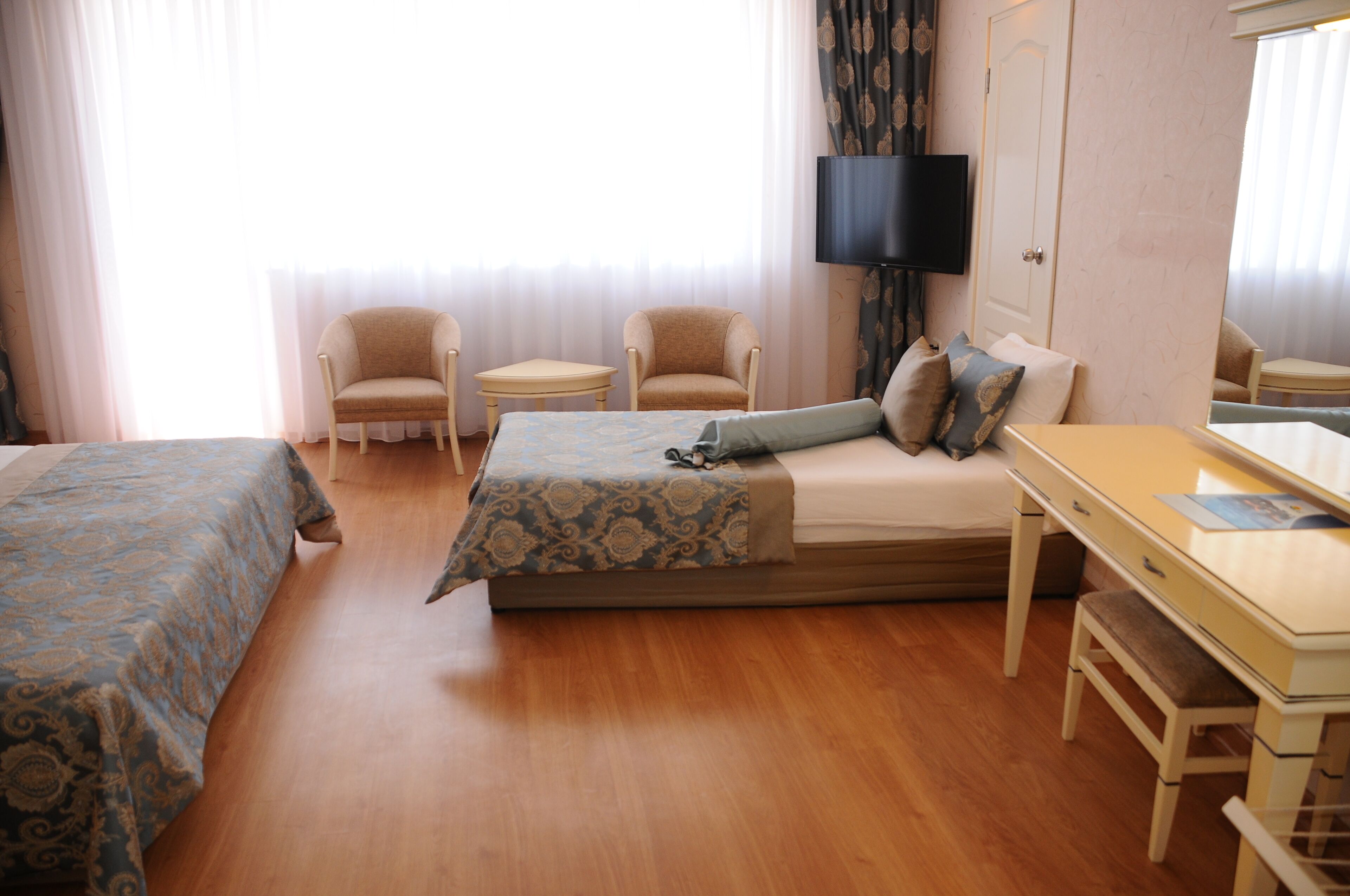 Quarto de hotel com cama, duas poltronas, uma mesinha, escrivaninha, televisão e janela com cortinas.
