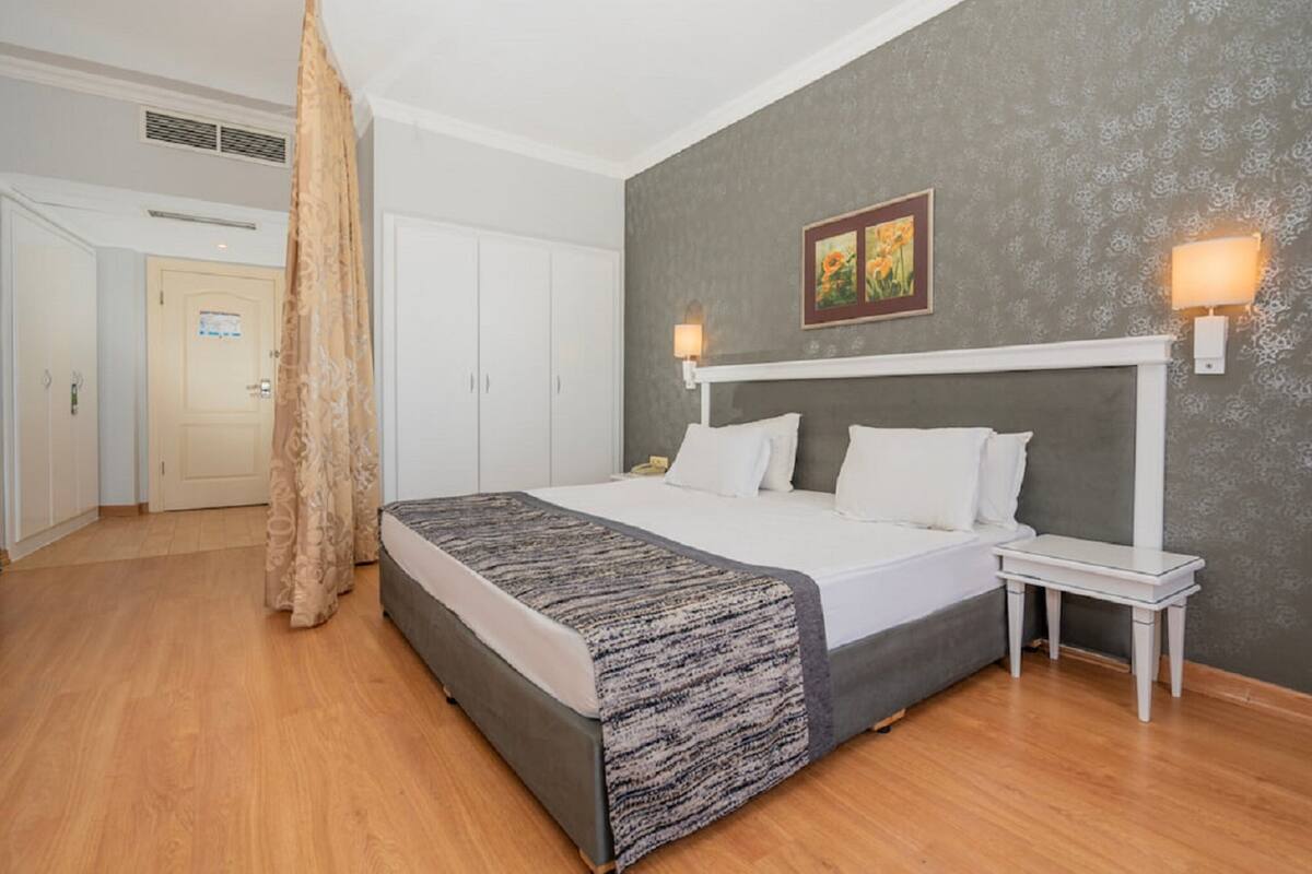 A hotel room with a large bed, a bedside table, a nightstand, a headboard, a wall-mounted lamp, and a door leading to another room.