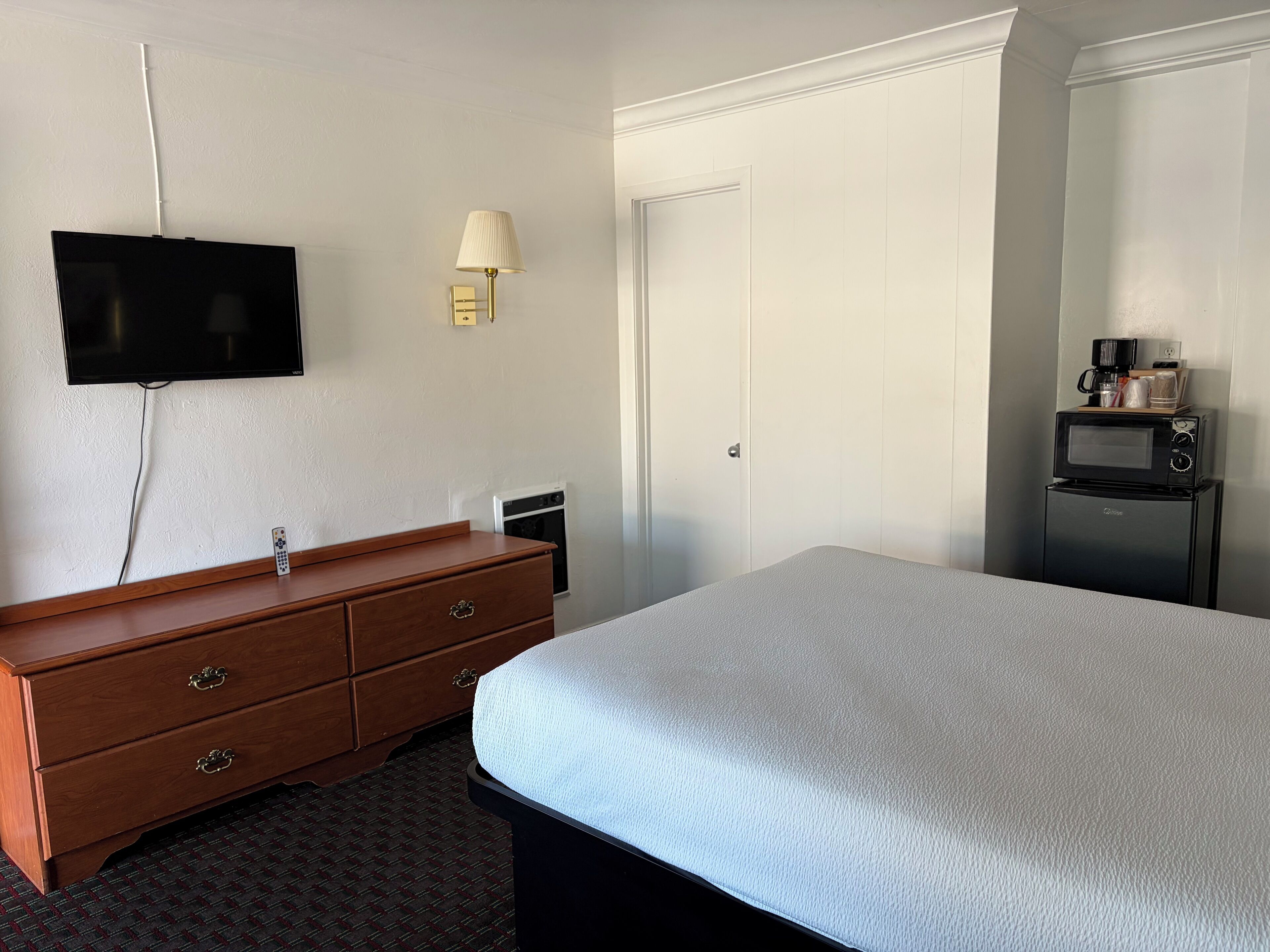 A hotel room with a bed, a dresser, a television mounted on the wall, a microwave, and a coffee maker on a small table.