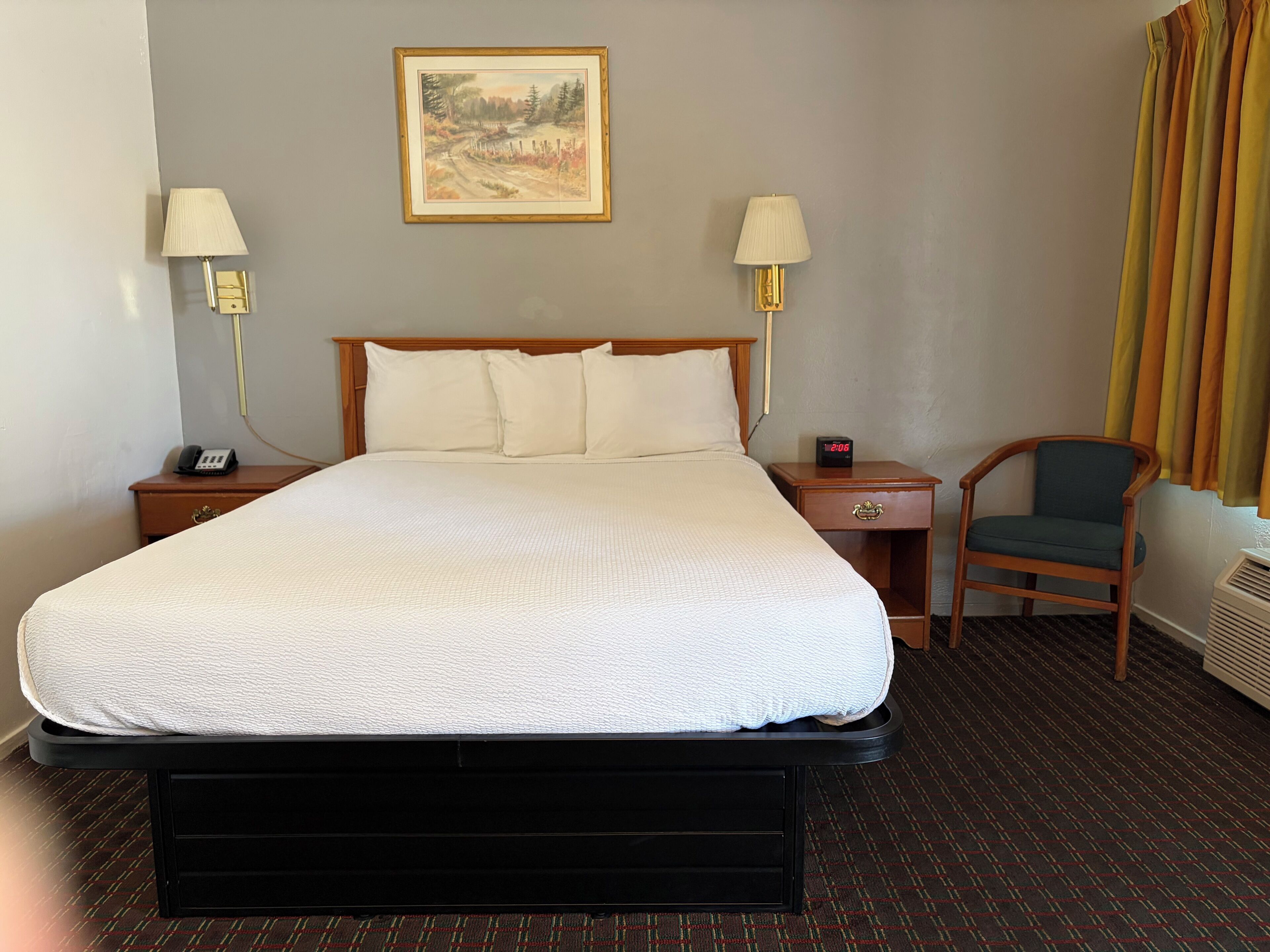 A hotel room with a bed, two bedside lamps, a nightstand with a phone, a chair, and a painting on the wall.