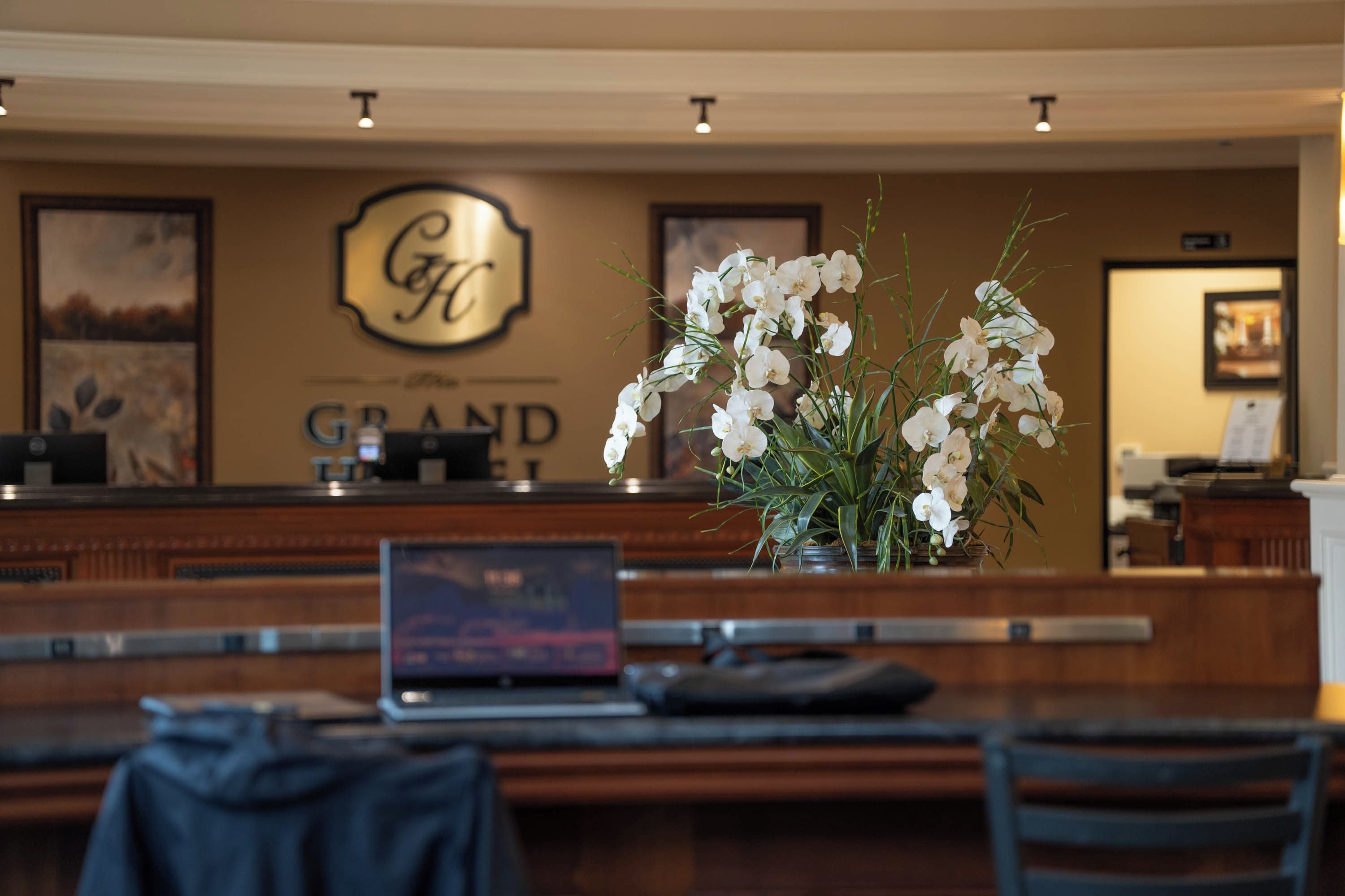 A hotel reception desk with a laptop, a vase of white flowers, and a coat hanging on a chair.
