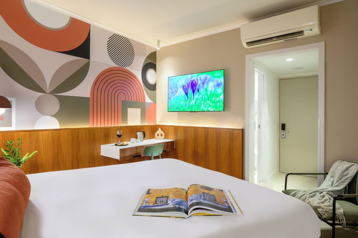 Una habitación de hotel moderna con una cama grande, un televisor en la pared, un escritorio con una silla y una puerta que da a otra habitación.