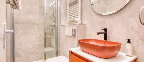 A modern bathroom with a glass shower enclosure, a white toilet, an orange vanity with a large bowl sink, and a round mirror on the wall.