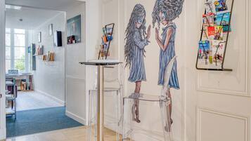 Un couloir avec une table en verre transparent, un mur orné d’une fresque et un panneau d’affichage où sont accrochés des magazines.