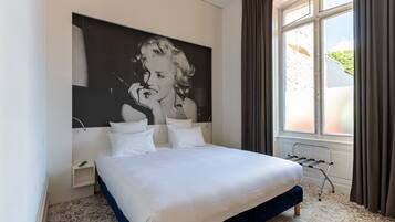 Un dormitorio moderno con una cama grande, una obra de arte colgada en la pared, una mesita de noche y una ventana con cortinas.