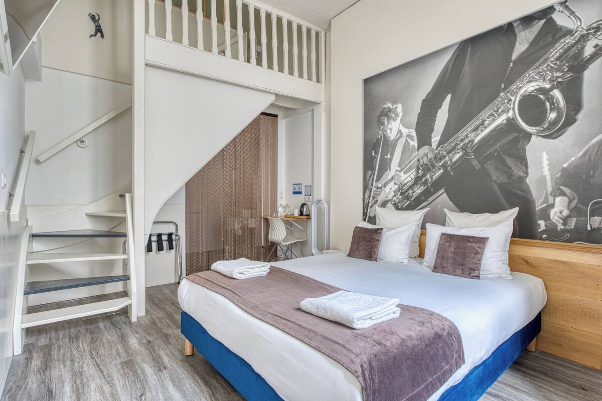 A modern hotel room with a large bed, a staircase, a desk, and a large poster of a saxophone player.
