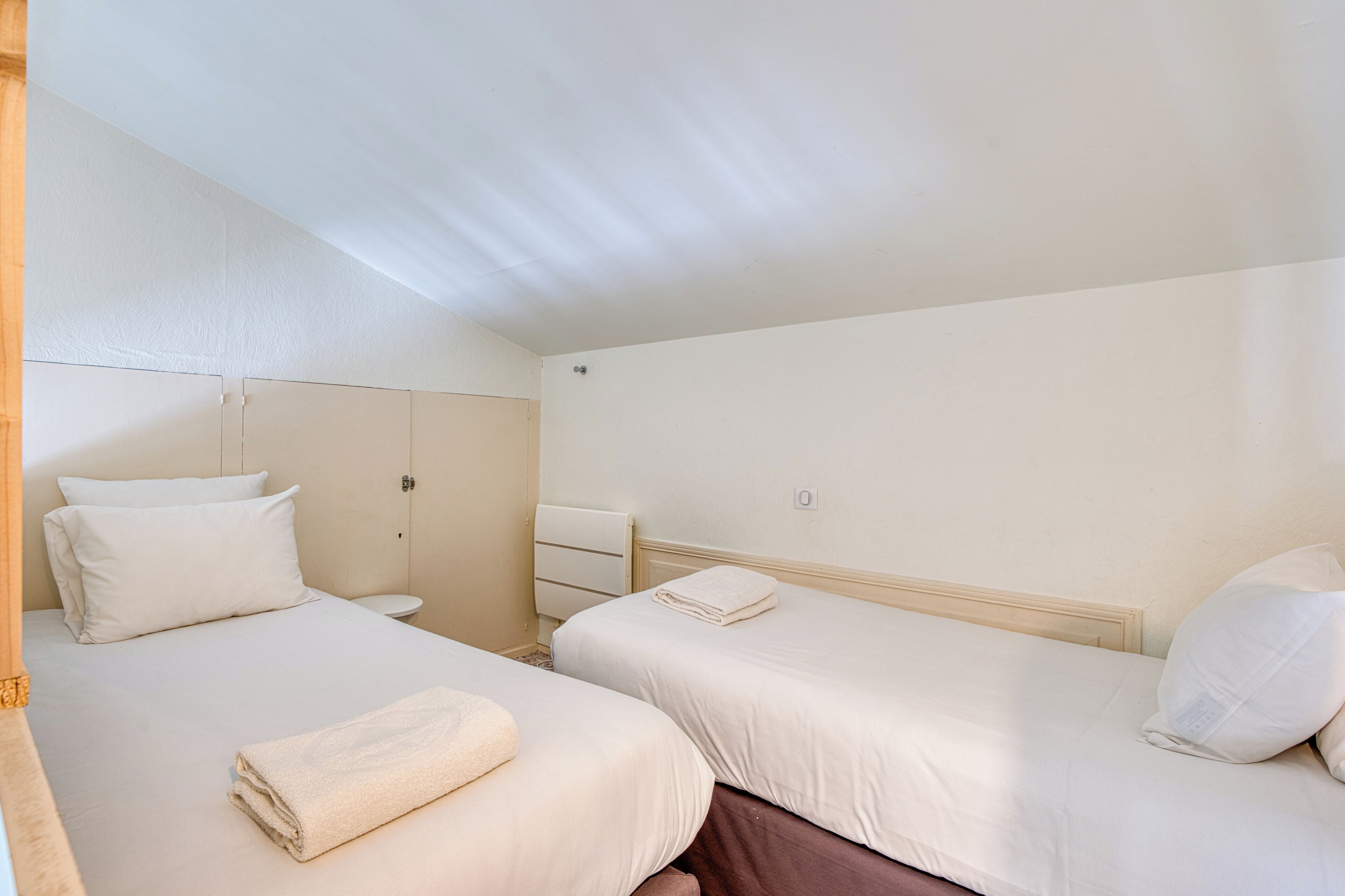 A hotel room with two beds, a small bedside table, a radiator, and a wall socket.