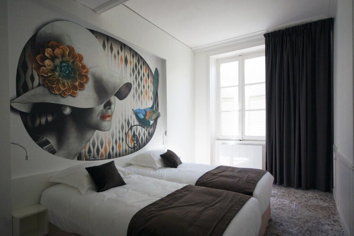 A hotel room with a large abstract painting on the wall, a bed with white and brown bedding, and a window with dark curtains.
