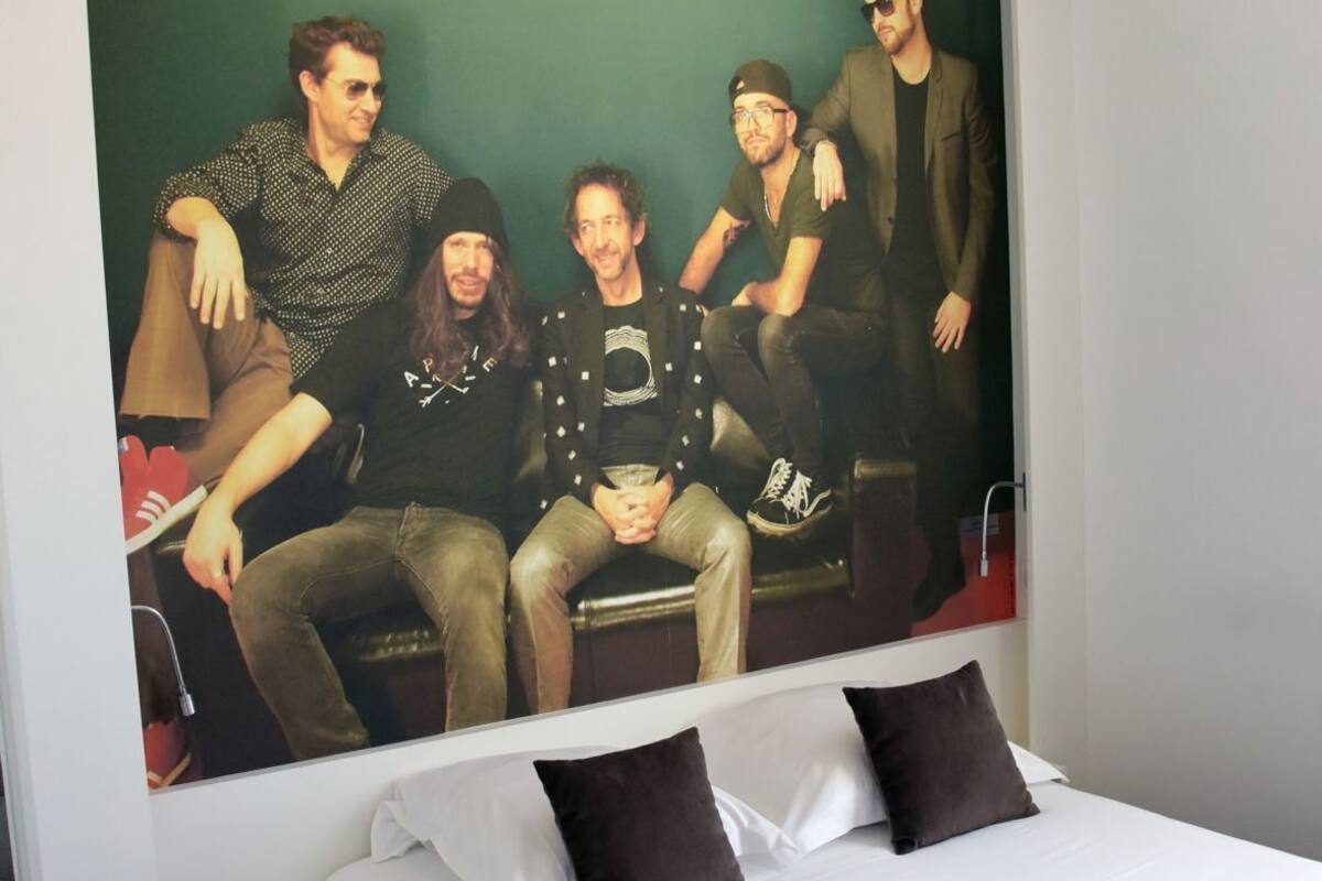 A hotel room with a large bed, a nightstand, and a wall-mounted painting of five people.