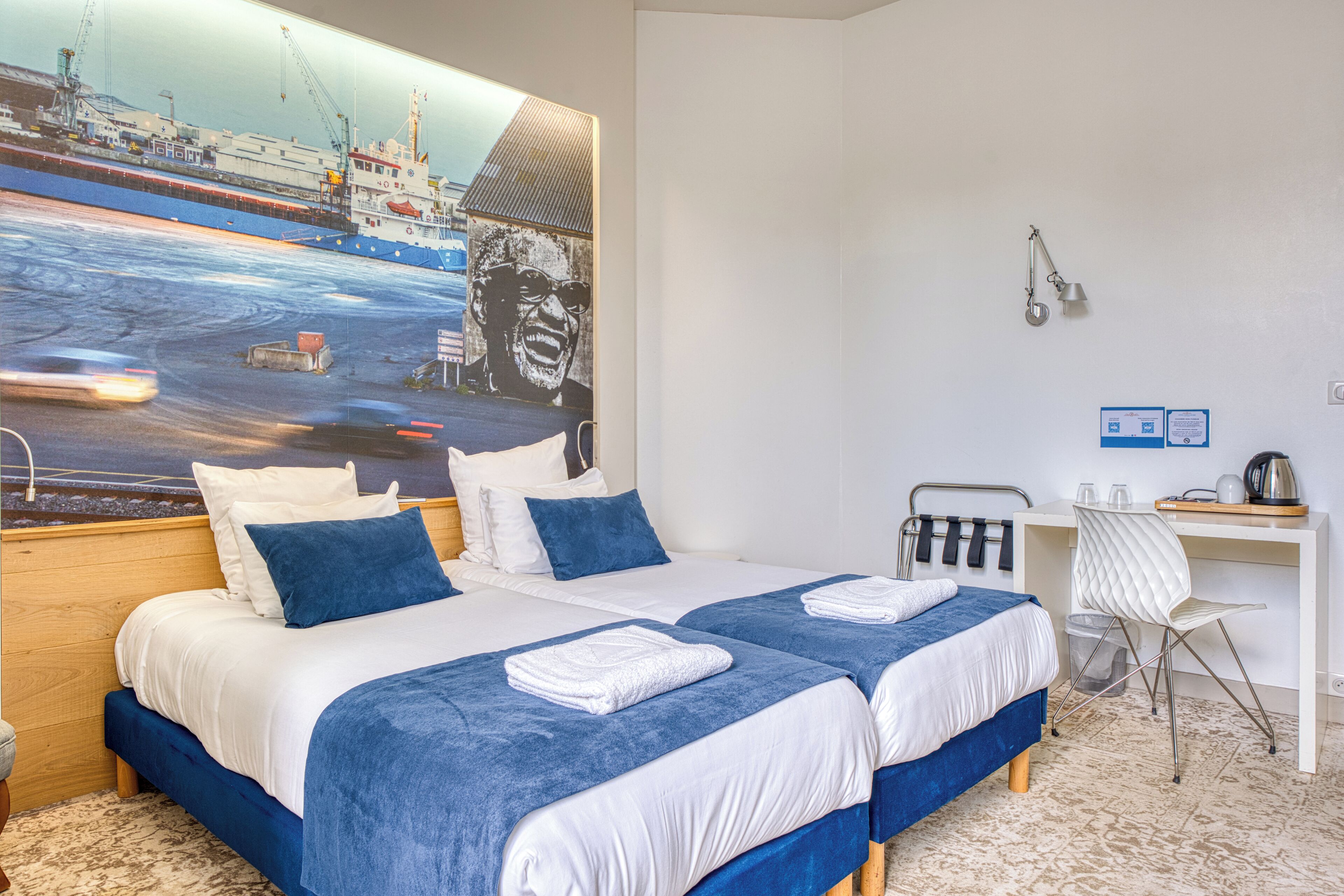 A hotel room with two beds, a desk, a chair, and a large painting of a ship on the wall.