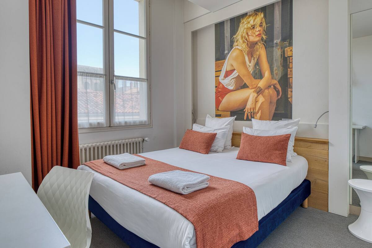 A hotel room with a large bed, a window with curtains, a painting on the wall, and a white bathroom.