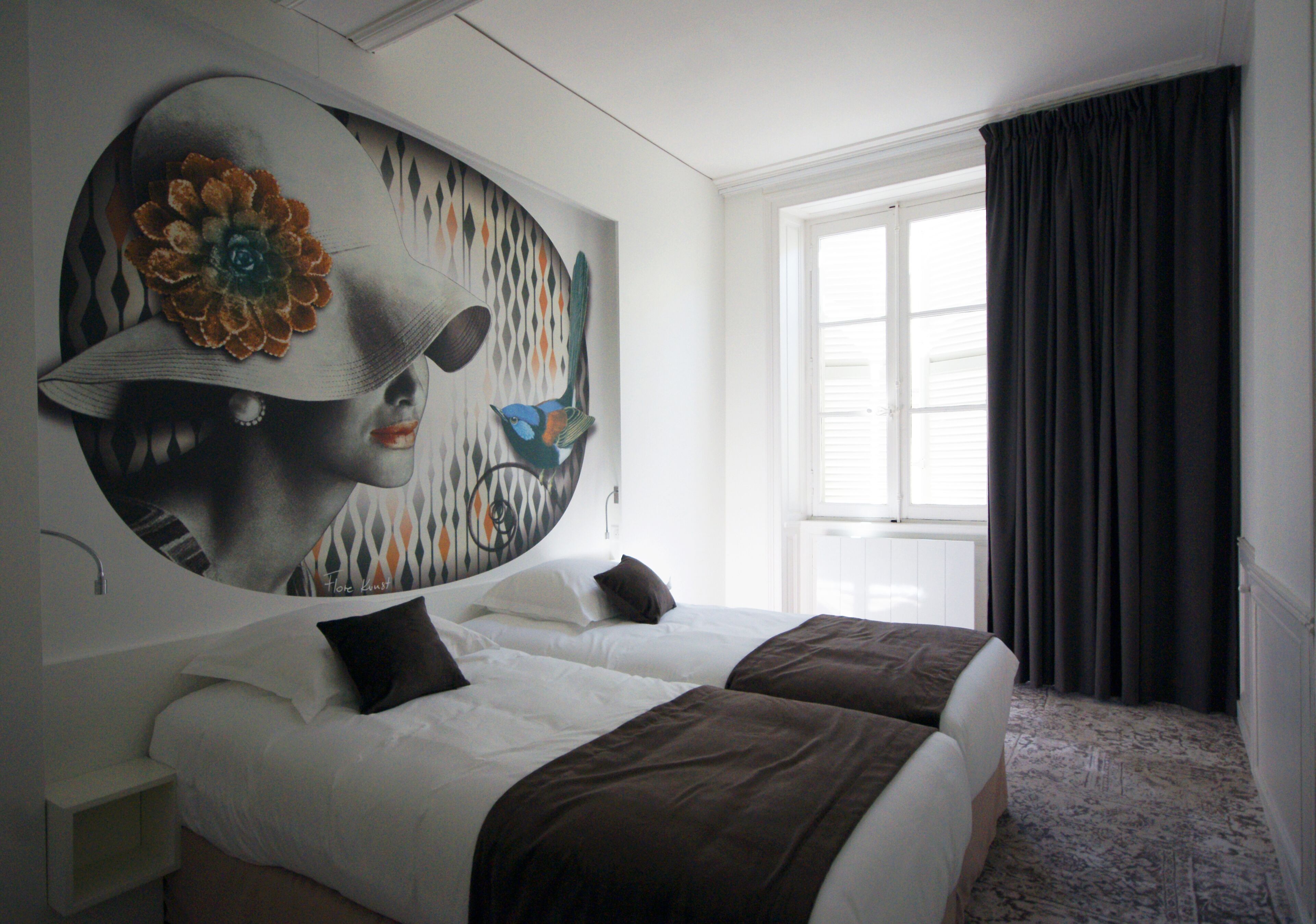 A hotel room with two beds, a large abstract painting on the wall, a window with dark curtains, and a carpeted floor.
