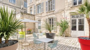 Une terrasse extérieure avec une table et des chaises, entourée de plantes en pot et dotée d’un sol pavé.