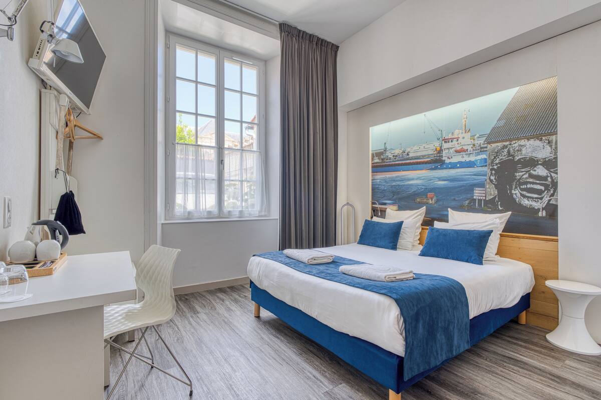 A modern hotel room with a large bed, a desk, a chair, and a large window.