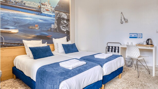 Une chambre d’hôtel avec deux lits, un bureau, une chaise et un grand tableau représentant un navire accroché au mur.