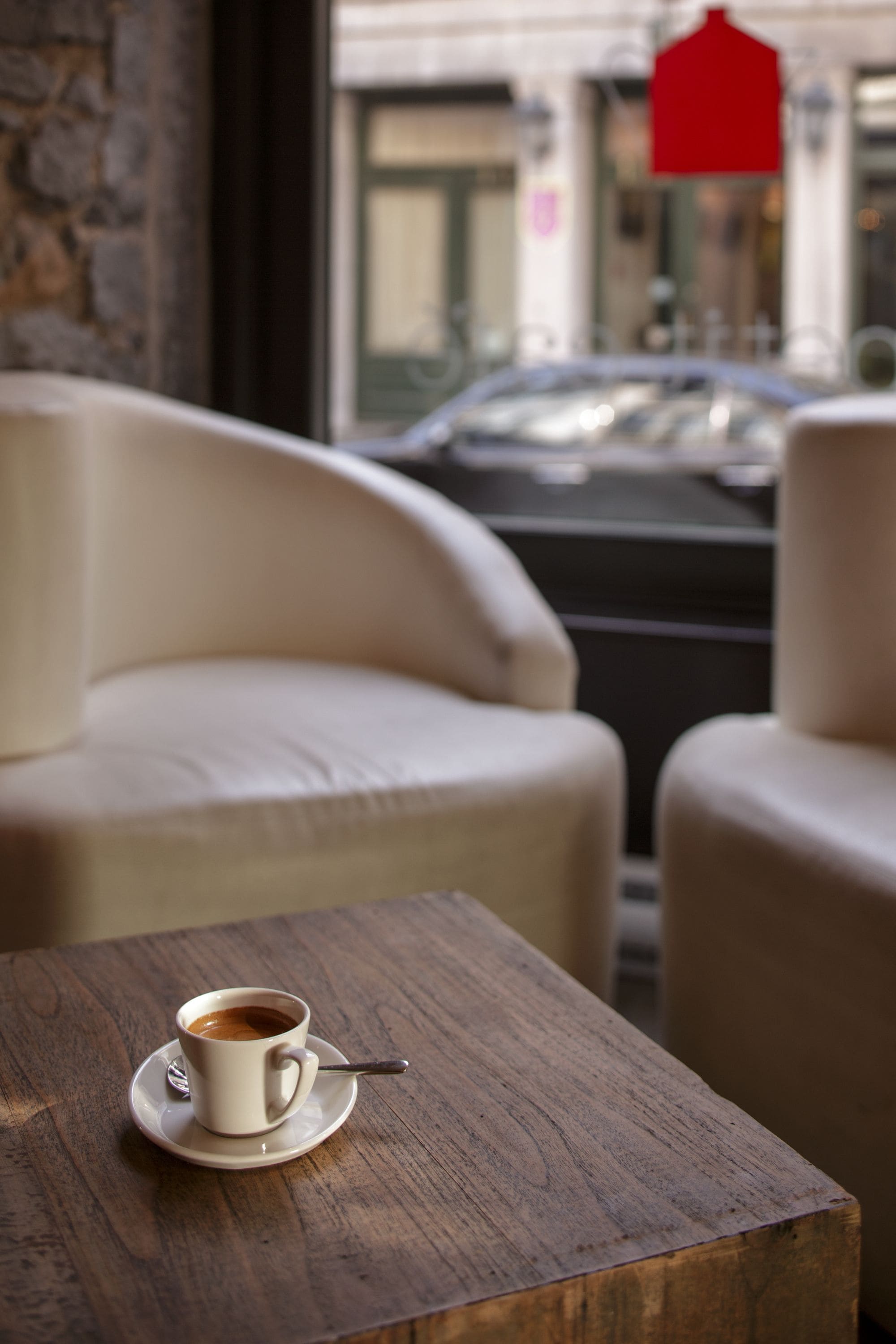 A cup of coffee on a wooden table in a cafe setting with a stone wall and a car visible outside.