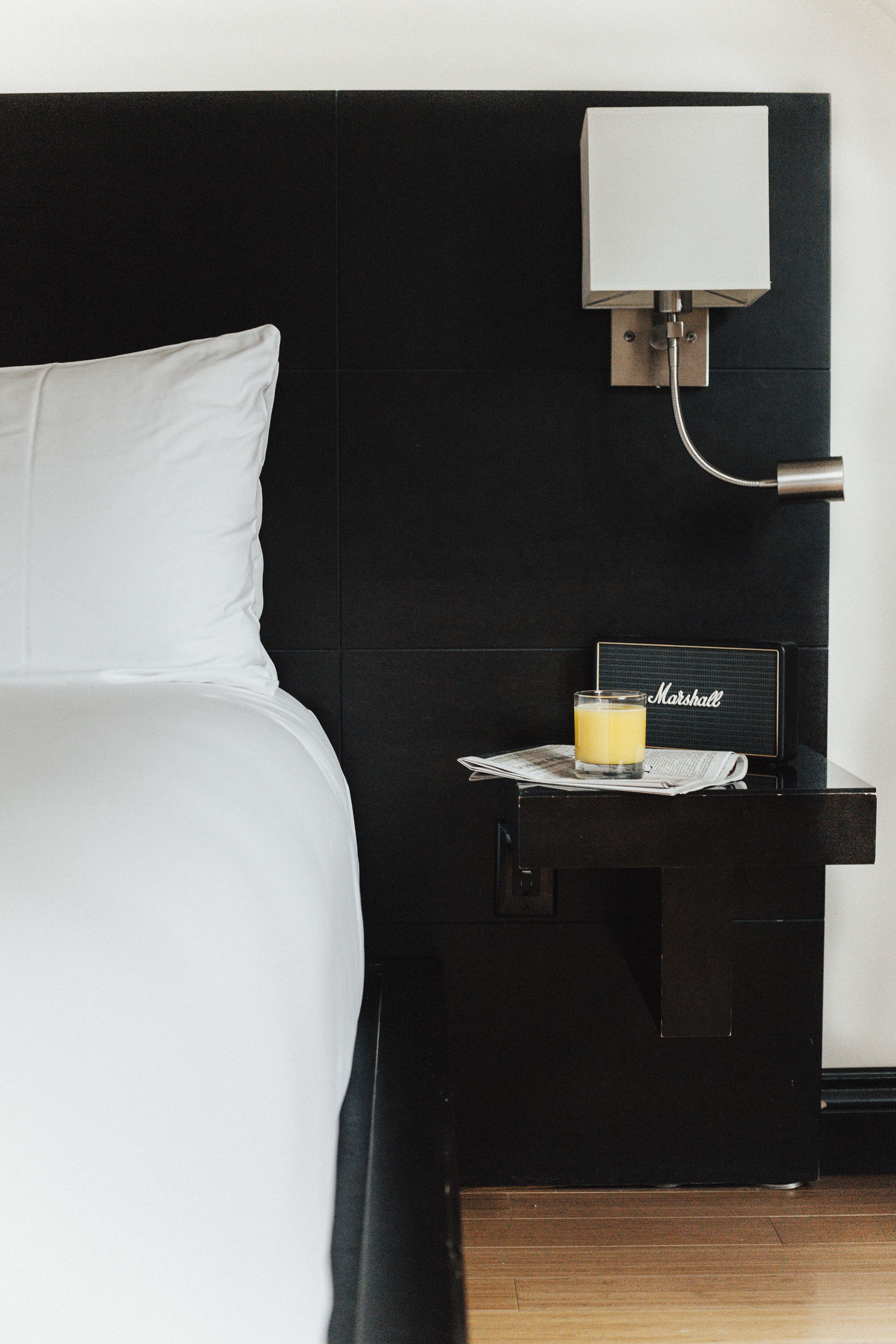 Un lit bien fait, avec du linge de lit blanc, une table de chevet sur laquelle se trouve un verre de jus d’orange, une lampe et une pancarte indiquant le nom « Marshall ».