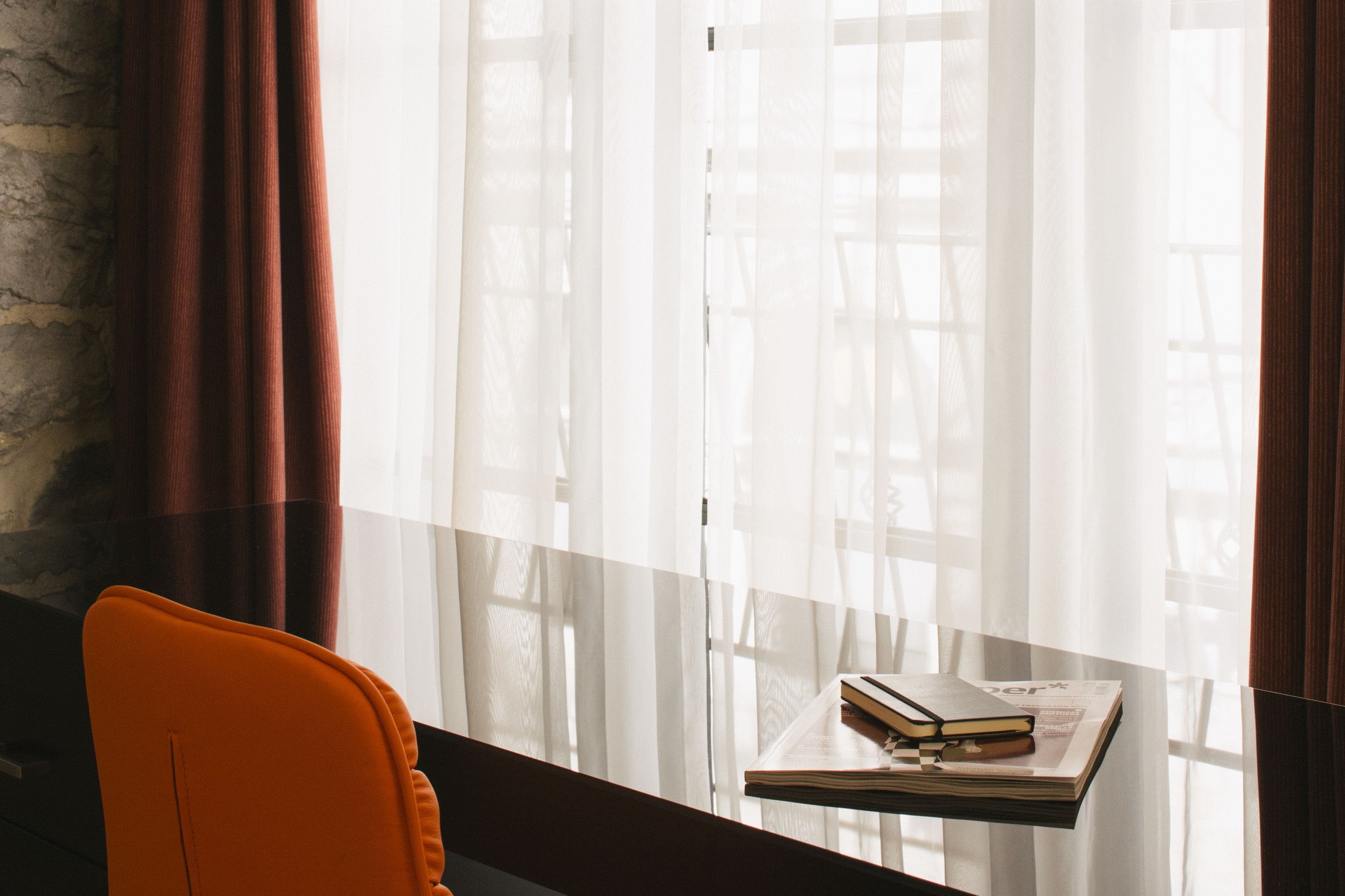 A room with a glass table holding a book and a magazine, a chair, and sheer curtains with a view of a building outside.