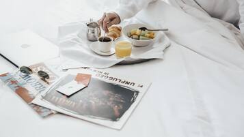Una bandeja de desayuno ordenada con un croissant, café, jugo y un cuenco de fruta sobre un edredón blanco.