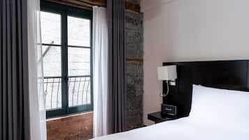 A hotel room with a bed, a nightstand with a lamp, a window with curtains, and a stone wall.