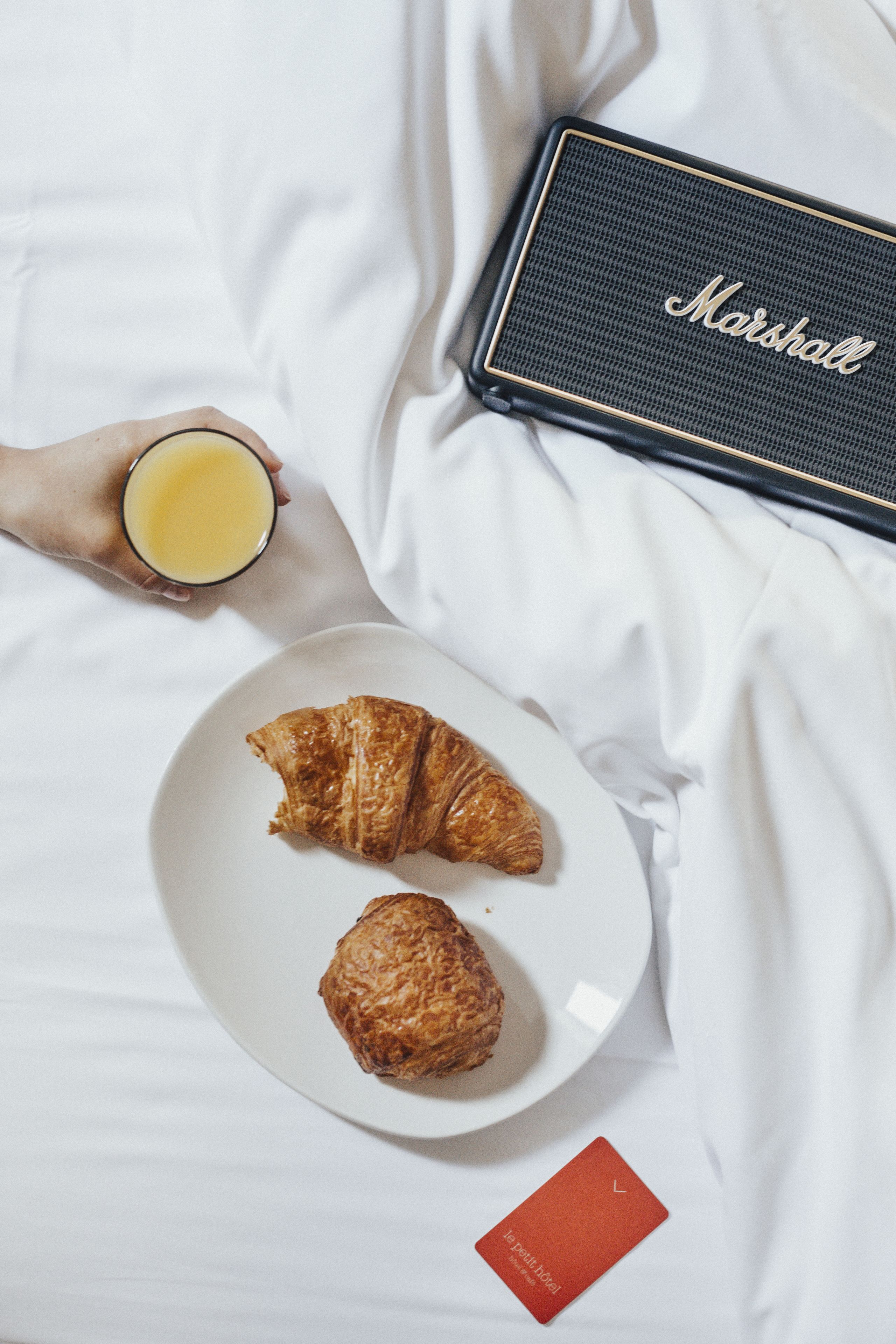 Ein Bett mit weißer Bettdecke, ein Teller mit zwei Croissants, ein Glas Orangensaft, ein Marshall Lautsprecher und eine rote Karte.