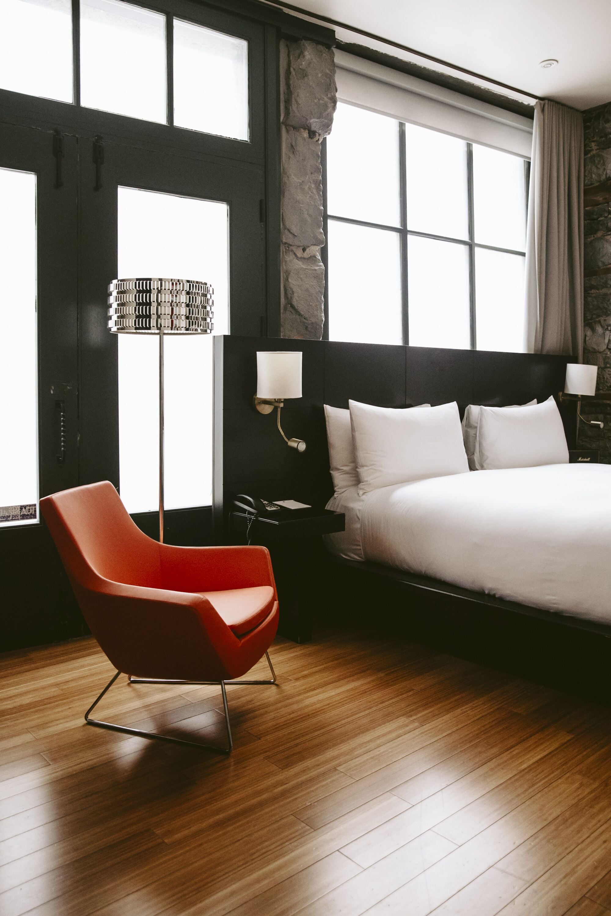 Ett modernt hotellrum med en stor säng, en röd fåtölj, en golvlampa och en vägg som har en dekorativ stenbeklädnad.