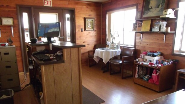 A wooden interior with a reception desk, a computer, a small table with chairs, and a shelf with various items. - Narrow Gauge Railroad Inn (Antonito)