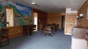 A room with wooden walls and a mural depicting a landscape. There are wooden tables and chairs, a refrigerator, and a ceiling fan. - Narrow Gauge Railroad Inn (Antonito)