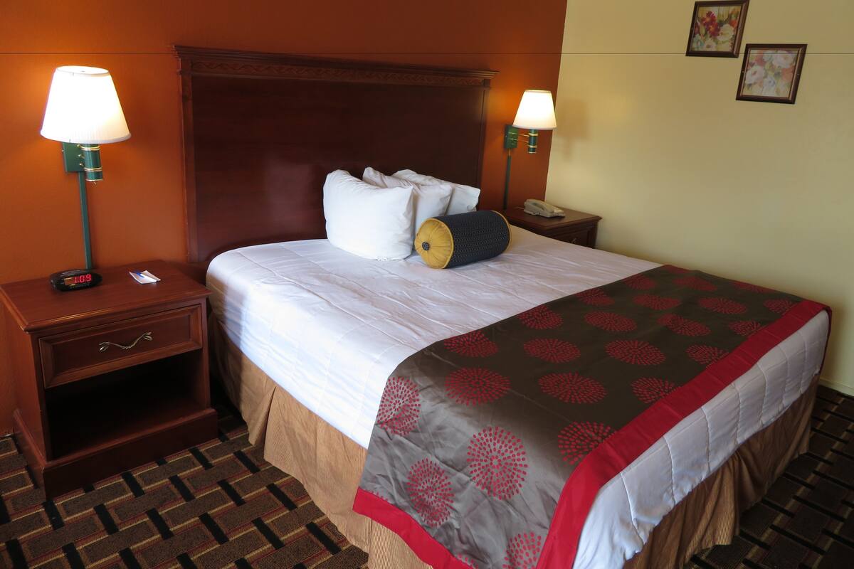 A hotel room with a large bed, two bedside lamps, a nightstand with a clock, a telephone, and a patterned carpet.