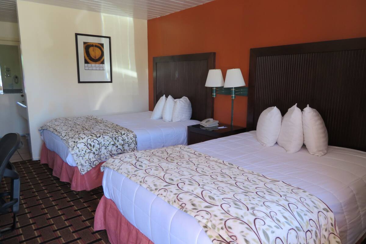 A hotel room with two beds, a desk, a chair, and a wall-mounted lamp.