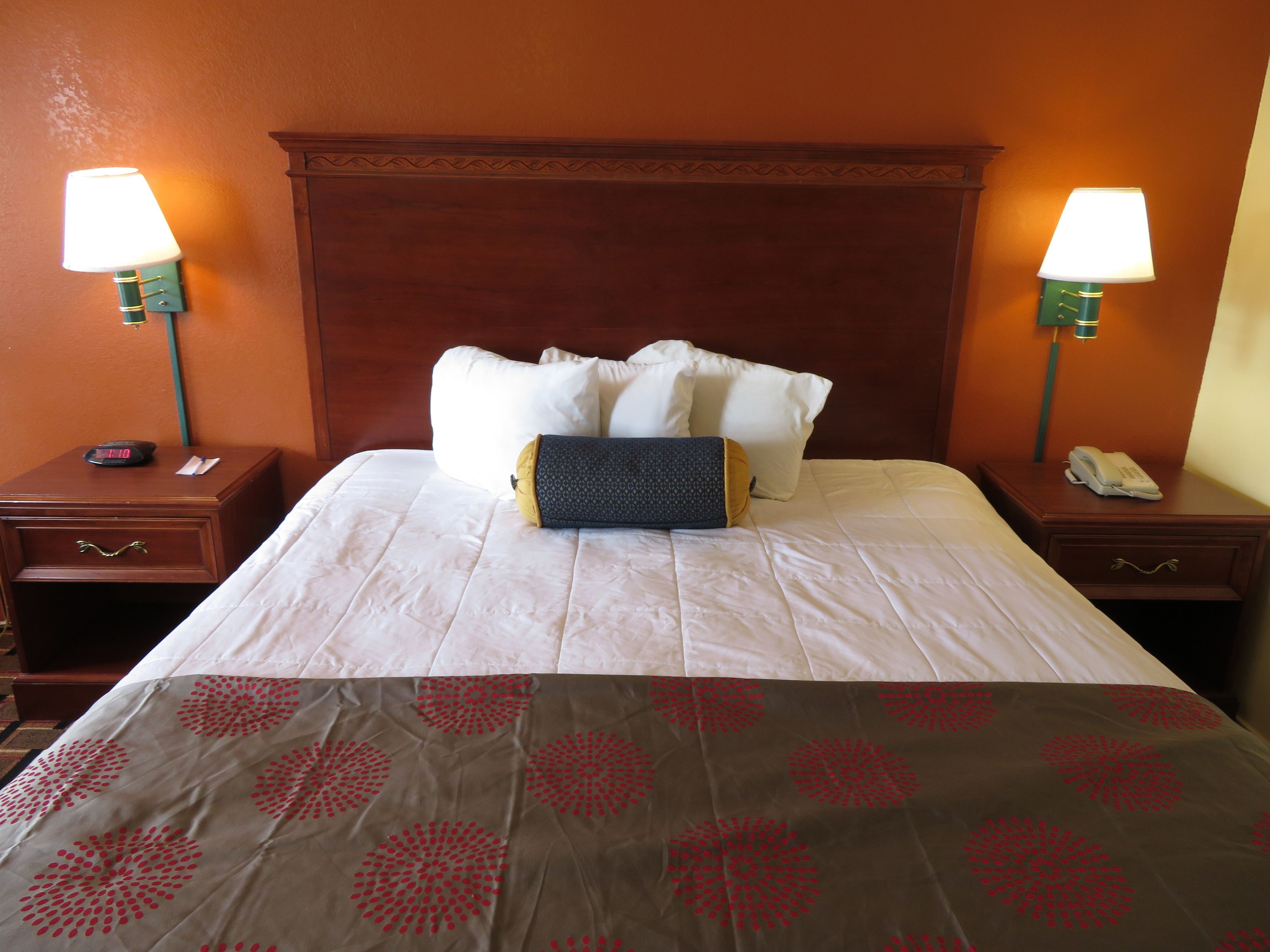 A hotel room with a neatly made bed, two bedside tables with lamps, a clock, and a phone on one of the tables.