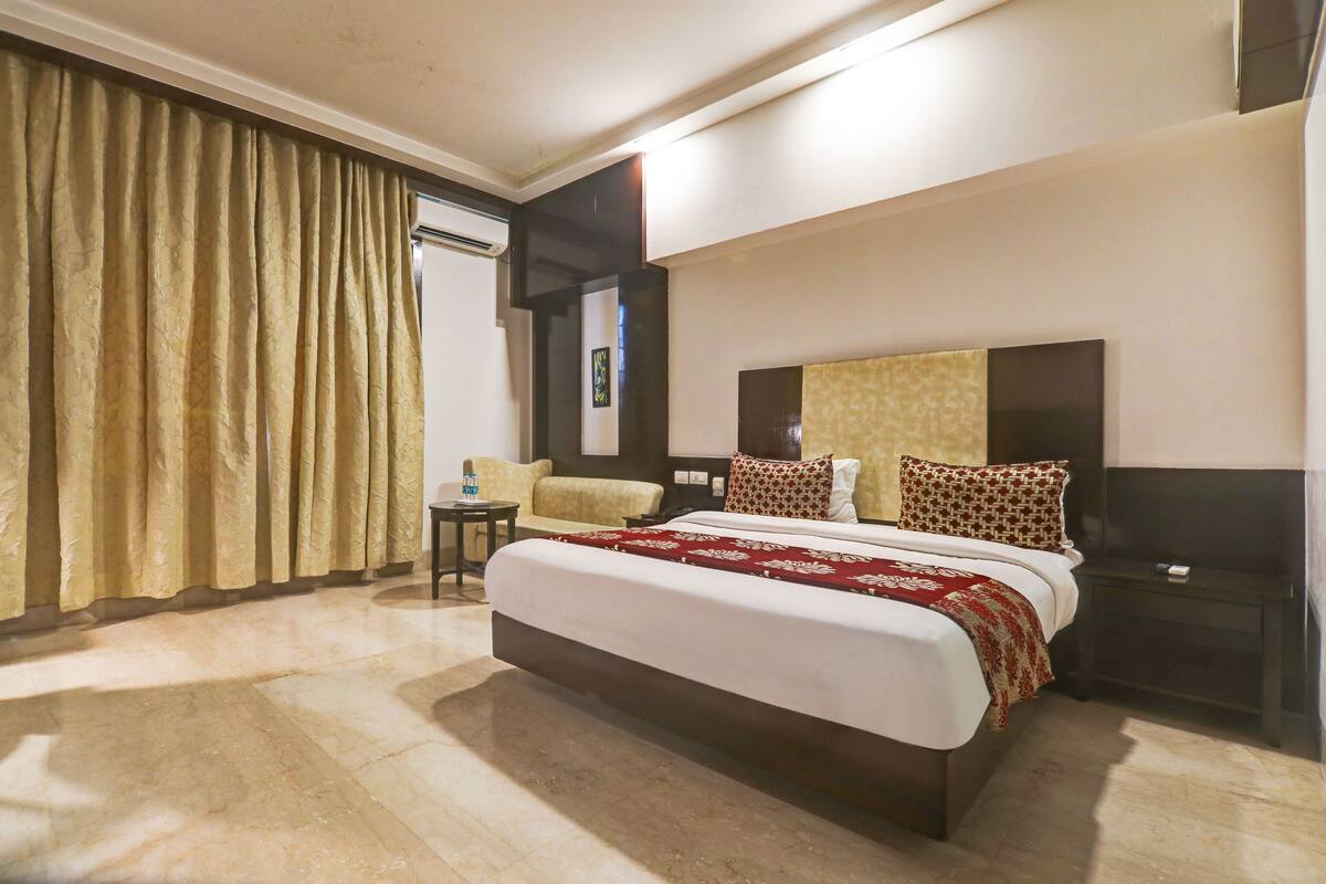 Une chambre d’hôtel avec un grand lit, une table de chevet, une commode, une chaise et une fenêtre avec des rideaux.